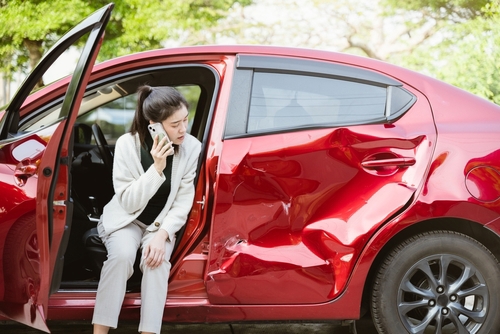 woman on phone sitting in open door of crash-damaged vehicle
